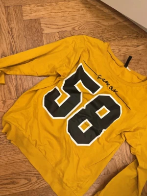 Gul långärmad topp med tryck - Cool gul långärmad topp med stort svart och vitt tryck av siffran 58 och texten 'Game On' på framsidan. Perfekt för dig som gillar sportig stil och vill sticka ut. Tillverkad i mjuk bomull för skön känsla hela dagen.