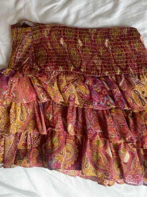 Mönstrad volangkjol i gult och rött - Kort volangkjol med paisleyinspirerat mönster i rött, gult och inslag av grönt. Kjolen har flera lager volanger och smock i midjan för en bekväm passform. Tillverkad i ett lätt, luftigt material som passar perfekt till varmare dagar.