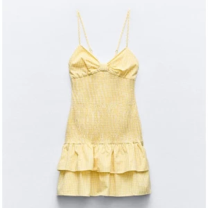 Gul rutig kort klänning med volang - Söt gul kort klänning med rutigt mönster och smala axelband. Klänningen har en volang längst ner och en lätt rynkad detalj vid bysten. Perfekt för varma dagar och somriga tillfällen.