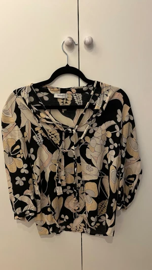 Blommig blus - Snygg blus från Max & Co med svart botten och beige blommönster. Blusen har trekvartsärm med resår vid ärmslut och dekorativ knytning vid halsen. Luftigt material som känns skönt mot huden och passar perfekt till jeans eller kjol.