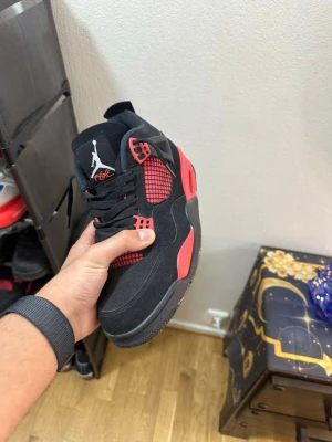 Svarta och röda Air Jordan 4 sneakers - Säljer ett par Air Jordan 4 sneakers i svart och rött med klassisk Jumpman-logga på plösen. Skorna har meshdetaljer, mockaliknande material och coola plastdetaljer vid snörningen. Perfekta för dig som gillar streetwear och vill sticka ut.