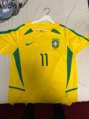 Brasiliens fotbollströja (Ronaldinho) t-shir - Den är för stor för mig de är därför jag säljer den. Gul Brasilien fotbollströja från Nike med gröna detaljer och nummer 11 på framsidan. Tröjan har korta ärmar, rund hals och är tillverkad i ett lätt, ventilerande material. Perfekt för fotboll eller som supporterplagg.