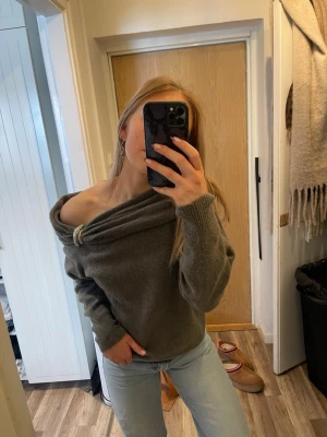 Grå off shoulder stickad tröja - Trendig grå stickad tröja med off shoulder-design och dekorativ metallring vid halsen. Mjuk och skön passform med långa ärmar och ribbade muddar. Perfekt för dig som vill ha en avslappnad men snygg look.