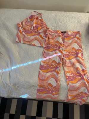 Färgglatt set med mönster - Säljer ett tvådelat set med ett linne och byxor i ett färgstarkt mönster med rosa, orange, vitt och lila. Linnet har smala axelband och byxorna är utsvängda i modellen. Perfekt för dig som gillar statement-plagg och vill sticka ut.