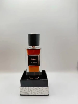 Ébène Eau de Parfum - Elegant parfymflaska i glas med mörk bärnstensfärgad vätska och svart etikett. Flaskan har en stilren, svart kork med textur och står på en dekorativ svart och silverfärgad låda. Perfekt för dig som gillar exklusiva dofter.