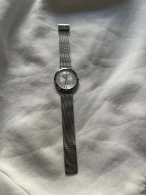 Silverfärgad klocka från Calvin Klein - Stilren klocka från Calvin Klein med rund urtavla i silver och minimalistisk design. Armbandet är i mesh och ger en modern känsla. Perfekt accessoar för dig som gillar enkel och tidlös stil. Skulle säga att det passar både herr/dam då urtavlan är relativt stor, men jag köpte den på damavdelning. Går att reglera passform. Batteribyte bör göras. Syns inte att den är använd. Frakten står köparen för :)