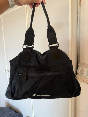 Svart handväska från Champion - Snygg svart handväska från Champion i sportig design. Väskan har dubbla handtag, en stor dragkedja upptill och en praktisk ytterficka med dragkedja framtill. Tillverkad i slitstarkt nylonmaterial med Champion-logga på framsidan.
