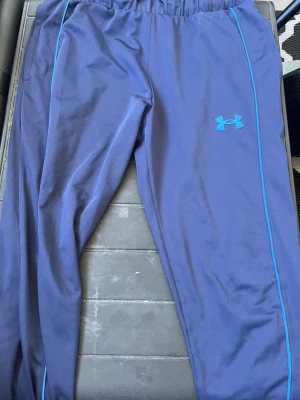 Blå träningsbyxor från Under Armour - Säljer ett par blå träningsbyxor från Under Armour med elastisk midja och ljusblå detaljer längs benen. Byxorna har en sportig look och är tillverkade i ett lätt och smidigt material som passar perfekt för träning eller chill.
