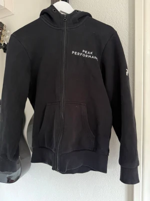 Svart hoodie Peak Performance - Snygg svart hoodie från Peak Performance med dragkedja framtill och fickor på magen. Märkets logga är tryckt på bröstet och ärmen. Ser ut som ny, använd ett par fåtal gånger