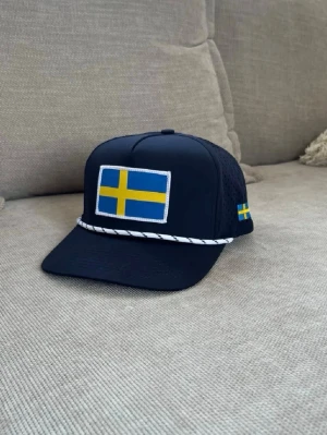 Mörkblå keps med svensk flagga - Snygg mörkblå keps från Golfio.se med broderad svensk flagga framtill och liten flagga på sidan. Kepsen har vit snodd över skärmen och ventilerande mesh på sidorna. Perfekt för dig som vill visa svensk stil på golfbanan eller i vardagen.