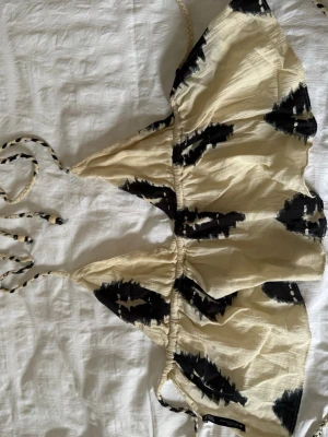Beige och svart topp från Zara - Säljer en beige topp från Zara med svarta abstrakta mönster. Toppen har tunna knytband i nacken och ryggen, vilket gör den enkel att justera. Materialet känns lätt och passar perfekt för strandhäng eller pool.