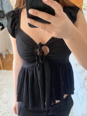 Svart blus med knyt och volang - Supersöt svart blus med knyt framtill och volangdetaljer vid axlarna. Toppen har en v-ringad urringning och ett litet hål under knytet som ger en extra touch. Luftigt fall och lätt rynkat tyg, perfekt för en trendig look.