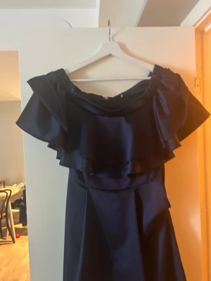 By Malina klänning, open shoulder med volang - Säljer en marinblå kortklänning från By Malina med stora volanger runt axlarna. Klänningen har en elegant offshoulder-look och är tillverkad i ett glansigt polyestermaterial som ger en lyxig känsla. Perfekt till bröllop!