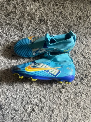 Blå Nike Zoom fotbollsskor - Säljer ett par blå Nike Zoom fotbollsskor med gula och vita detaljer samt snörningsfritt design. Skorna har högre skaft och mönstrad ovandel för extra grepp på planen. Sulan har gula dobbar för bra fäste på gräs. Perfekta för dig som vill sticka ut på planen.