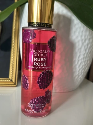 Victoria's Secret Ruby Rosé parfym - En snygg parfymmist från Victoria's Secret med doft av hallon och rosblad. Flaskan är transparent med rosa färg och dekorativa hallonmotiv, samt har en elegant guldfärgad kork. Perfekt för dig som gillar fruktiga och blommiga dofter.