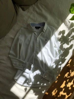 Vit skjorta från Ralph Lauren 100%bomull knit Oxford - Ny vit skjorta från Ralph Lauren med broderad logga på bröstet. Insidan av kragen har blåa ränder för detalj samt insidan av ärmen som syns när den är uppvikt. Tillverkad i mjuk bomull som är sval och rörlig, perfekt till sommaren. Storlek L men liten i storleken så passar m/s lika bra. Modell knit oxford