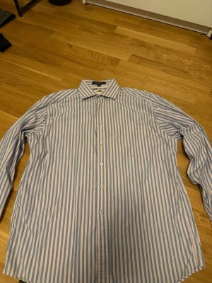Randig skjorta från Tommy Hilfiger - Säljer en klassisk skjorta från Tommy Hilfiger med ljusblå och vita vertikala ränder. Skjortan är tillverkad i 100% bomull och har en snygg krage med broderad logotyp på insidan. Perfekt för dig som gillar stilrena plagg.