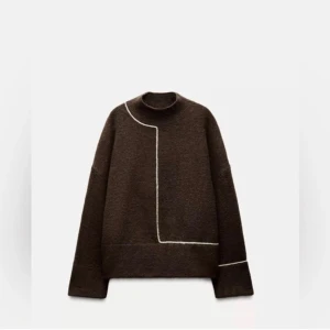 Populär stickad tröja från Zara  - Snygg brun stickad tröja med lång ärm och hög hals. Tröjan har en vit linje som går över axeln, ner på framsidan och runt ena ärmen vilket ger en cool grafisk look. Passformen är loose och materialet känns mjukt och varmt.
