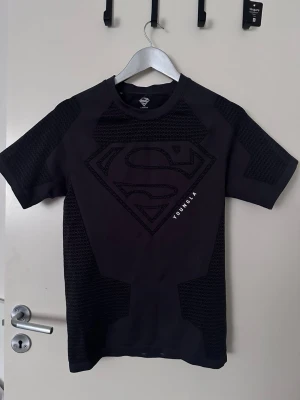 Svart t-shirt från YoungLA med Superman - Cool svart t-shirt från YoungLA med stort Superman-logotyp på bröstet. Materialet är en mix av slät och strukturerad textil som ger en sportig känsla. Perfekt för gymmet eller träning, med korta ärmar och rund hals.