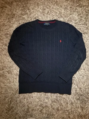 Mörkblå kabelstickad tröja Polo Ralph Lauren - Kabelstickad mörkblå tröja från Polo Ralph Lauren med rund halsringning och klassisk röd logga broderad på bröstet. Tröjan har långa ärmar och ribbade muddar vid ärmslut och nederkant. Perfekt för lager på lager och stilren look.