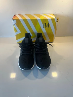 Svarta Adidas Ultraboost sneakers - Säljer ett par svarta Adidas Ultraboost sneakers med stickad mesh-överdel och klassiska tre ränder på sidan. Skorna har en vit Boost-sula som ger grym komfort och svart yttersula. Perfekt för dig som vill ha både stil och funktion.