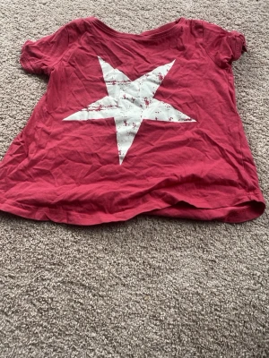 Röd t-shirt med vit stjärna - Cool röd t-shirt med en stor vit stjärna tryckt på framsidan. T-shirten har korta ärmar och en avslappnad passform. Perfekt för dig som vill sticka ut lite extra med en enkel men snygg detalj.