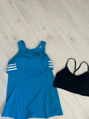 Blå Adidas sport-bh  & svart träningsbyxor - Säljer en blå Sportbhfrån Adidas med vita ränder på sidorna och meshdetaljer i ryggen. Tillhörande svart sport-bh med tunna axelband. Perfekt för  träning och löpning. Materialet är mjukt och stretchigt.