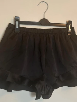 Shorts - Svarta korta shorts i satin med resår i midjan. I mycket fint skick. Volanger i benöppningarna. Material: Polyester