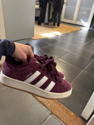 Adidas Campus vinröda sneakers - Vinröda Adidas Campus sneakers med klassiska vita ränder på sidan och vit sula. Skorna har en låg profil och är tillverkade i mocka med matchande vinröda skosnören. Perfekt för dig som vill ha en stilren och cool look.