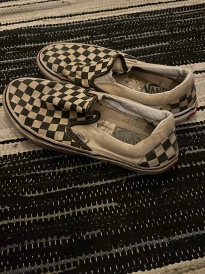 Svartvita checkerboard Vans slip-on - Klassiska Vans slip-on sneakers med svartvitt checkerboard-mönster. Låg modell utan snörning och vit sula med svart rand. Ovandelen är i canvas och insidan är vadderad för extra komfort. Perfekta för en avslappnad streetwear-look.