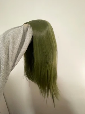 Lång grön hårperuk - Säljer en lång och rak hårperuk i en cool olivgrön nyans. Peruken har naturlig glans och är tillverkad av syntetiskt material som känns mjukt och följsamt. Perfekt för dig som vill testa en ny look eller sticka ut med färgstarkt hår.