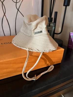 Beige bucket hatt  - Cool beige bucket hat  med rå kant och justerbart band under hakan. Hatten är tillverkad i bomull och har en metallogo på sidan. Perfekt för att lyfta din streetstyle-look.