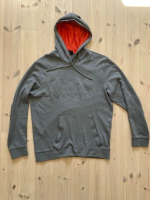 Grå hoodie från Hugo Boss - Säljer en grå hoodie från Hugo Boss med broderad logga på bröstet. Hoodien har en huva med orange foder och dragsnören samt en stor magficka. Materialet är mjukt och bekvämt. I bra skick och inga defekter. 