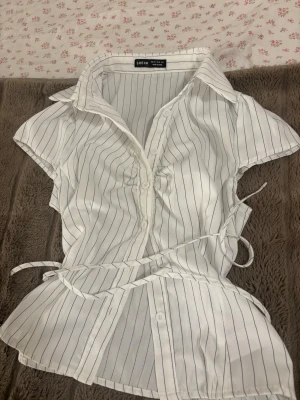 Vit randig blus från SHEIN - Snygg vit blus från SHEIN med tunna svarta ränder, knappar framtill och ett matchande midjeband. Blusen har krage och korta ärmar, perfekt för en trendig och fräsch look. Tillverkad i ett lätt och luftigt material. 