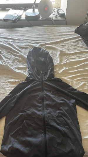 EA7 tröja - Säljer en svart hoodie med dragkedja och stora fickor framtill. Hoodien har en rymlig huva och är tillverkad i ett mjukt material som känns bekvämt. Perfekt för dig som gillar en enkel och stilren look.