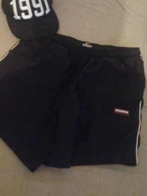 Svarta shorts från McKenzie och en keps  - Svarta shorts från McKenzie med vita detaljer längs sidorna och logotyp på ena benet. De har elastisk midja och sportig design, perfekta för en avslappnad stil. Materialet känns lätt och bekvämt, idealiskt för varma dagar.