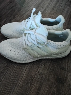 Vita Adidas Ultraboost sneakers - Snygga vita Adidas Ultraboost sneakers med stickad mesh-överdel och klassiska tre ränder på sidan. Skorna har en tjock och bekväm Boost-sula samt vita skosnören. Perfekta för dig som gillar sportig stil och komfort.