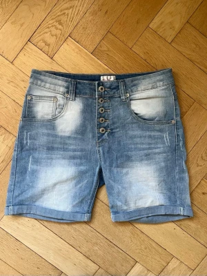 Blå denimshorts med knappar - Säljer ett par blå denimshorts med fem knappar framtill och klassiska fickor både fram och bak. Shortsens tvättade look ger en cool och avslappnad känsla. Mycket stretchight material!