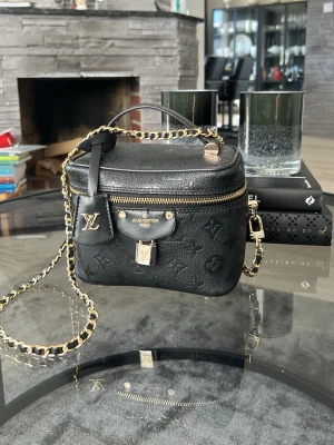 Svart handväska - Säljer en exklusiv svart handväska. Väskan har guldfärgade detaljer, kedjerem och ett elegant lås framtill. Perfekt för dig som vill ha en lyxig och trendig accessoar.