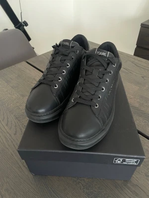 Svarta sneakers från Karl Lagerfeld - Snygga svarta sneakers från Karl Lagerfeld med diskret logotyp på sidan och mönstrad yta. Skorna har svarta skosnören, metallfärgade öljetter och är tillverkade i läder med en klassisk låg profil. Perfekt för dig som gillar stilrena och trendiga skor.