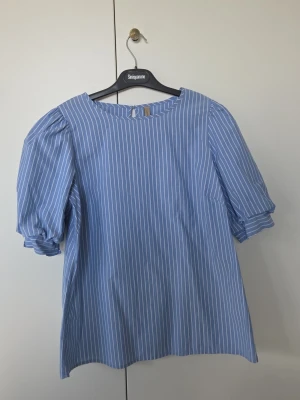 Blåvit randig blus med puffärm - Blus i ljusblått med vita vertikala ränder och puffiga korta ärmar. Rund halsringning och knapp i nacken. Luftig och bekväm, perfekt för en trendig och avslappnad look. Passar till både jeans och kjol.