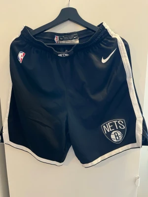 Brooklyn Nets basketshorts Nike - Säljer ett par svarta basketshorts från Nike med Brooklyn Nets-logga och vita detaljer längs sidorna. Shortsens material är lätt och ventilerande mesh, perfekt för träning eller match. NBA-logga och elastisk midja för skön passform. Storlek ~M 