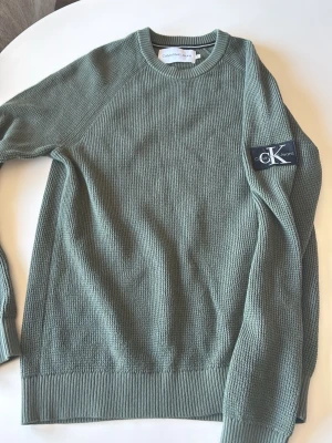 Olivgrön stickad tröja från Calvin Klein - Säljer en olivgrön stickad tröja från Calvin Klein med rund halsringning och ribbade muddar. Tröjan har ett diskret CK patch på ena ärmen och är tillverkad i mjukt material. Storlek M passar perfekt för den som är mellan 175-185 ca.     Tröjan har bara hängt i min garderob utan användning, därför den säljs. Tröjan är som sagt helt ny.
