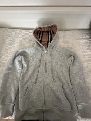 Grå BB zipper  - Snygg grå zip hoodie från BB med klassiskt rutigt foder i huvan och broderad logga på bröstet. Hoodien har dragsko i huvan, två stora fickor fram och ribbade muddar. Perfekt för en chill och stilren look. Det står M size men de är S och XS