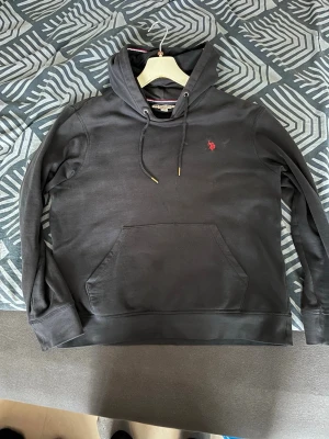 Marinblå hoodie från U.S. Polo Assn - Snygg svart hoodie från U.S. Polo Assn med klassisk känguruficka och dragsko i huvan. Liten röd logga broderad på bröstet. Tillverkad i mjukt material som känns skönt mot huden. Perfekt för en avslappnad och stilren look.