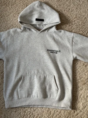 Grå Essentials hoodie  (Fear of God) - Säljer en grå hoodie från Fear of God Essentials med svart logotyp på bröstet och ryggen. Hoodien har en stor huva, ribbade muddar och en klassisk magficka. Materialet är mjukt och bekvämt, perfekt för en avslappnad look. I storleken small, bästa kvaliteten.