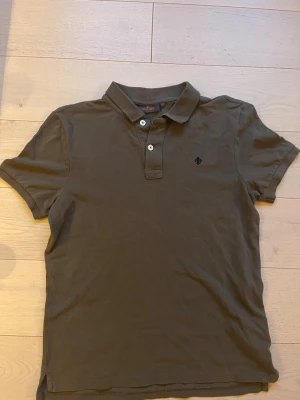 Olivgrön pikétröja från Morris - Snygg olivgrön pikétröja från Morris med klassisk krage och två knappar framtill. Tröjan har korta ärmar och en diskret broderad logga på bröstet. Tillverkad i bomull.