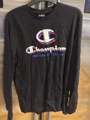 Svart långärmad topp från Champion - Svart långärmad topp från Champion med stort broderat logotyptryck i vitt, blått och rött på bröstet. Rund halsringning och normal passform. Perfekt för dig som gillar sportiga och klassiska streetwear-vibes.