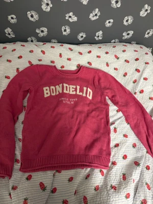 Rosa stickad tröja från Bondelid - Rosa stickad tröja från Bondelid med vit text på bröstet och rund halsringning. Tröjan har ribbstickade muddar vid ärmslut och nederkant, samt långärmad design. Perfekt för dig som gillar klassisk och enkel stil.
