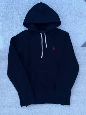 Svart hoodie från Polo Ralph Lauren - Svart hoodie med vit dragsko och klassisk röd Polo-logga broderad på bröstet. Hoodien har en stor huva och en praktisk magficka. Perfekt för dig som gillar stilren design och ikoniska märken. Som ny